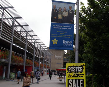 Brunelin kampuksella opiskelijoita ja lyhtypylväässä kirkasvärinen julistemyynti-ilmoitus sekä yliopiston kansallisesta menestyksestä kertova juliste